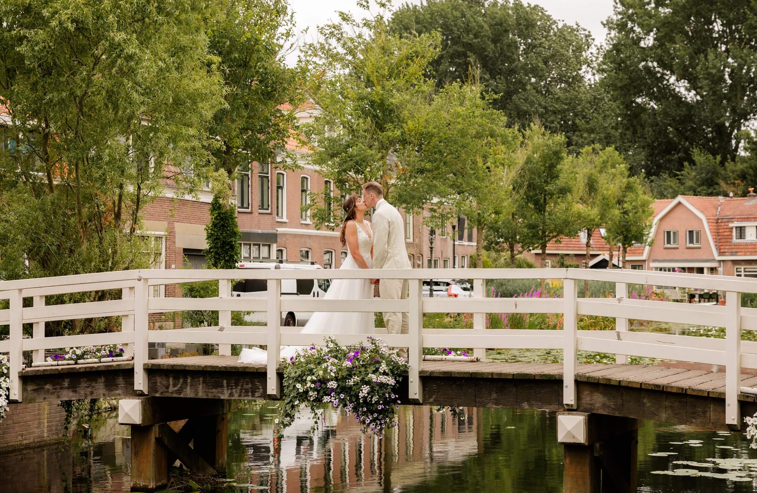 Bruidspaar zoent elkaar op een witte brug over gracht in Weesp, omringd door groen en huizen