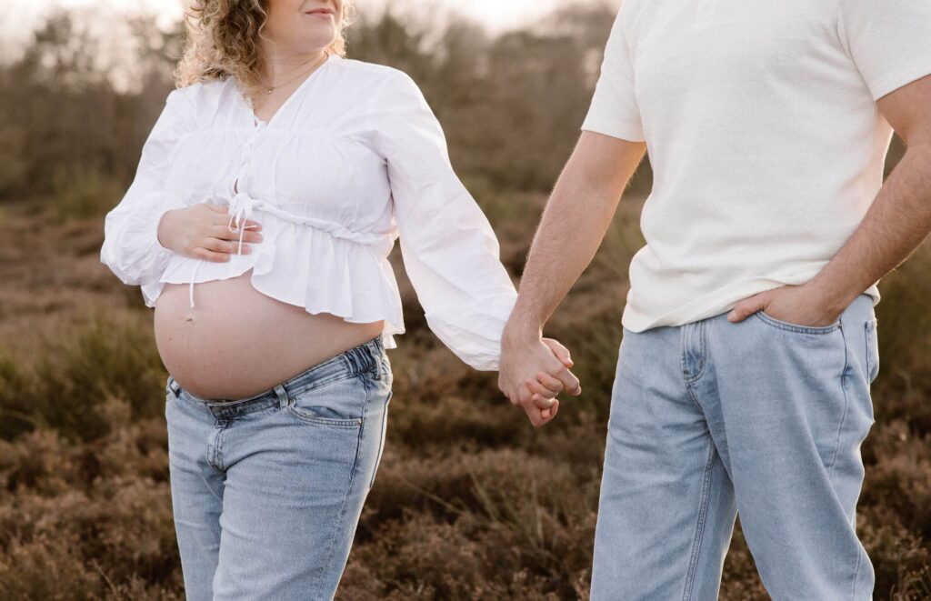 Zwangere vrouw in witte blouse en jeansbroek houdt handen vast met haar man in witte t-shirt en jeansbroek tijdens zwangerschapsshoot in het bos