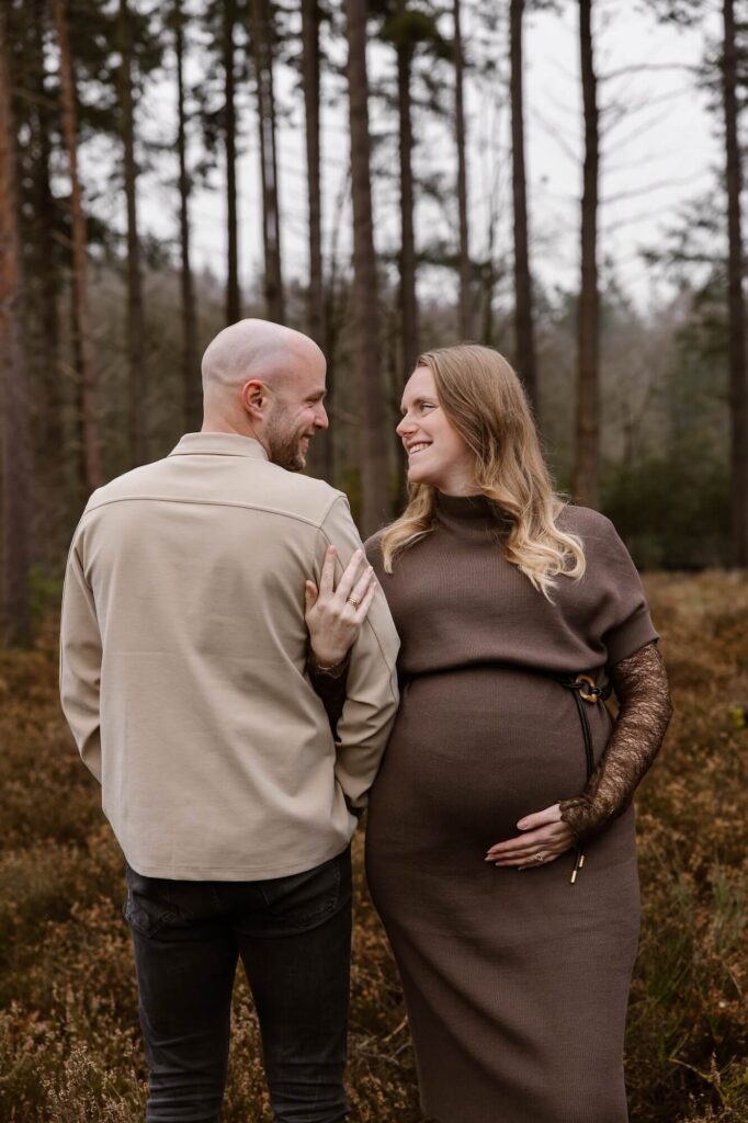 Zwangere vrouw in bruine jurk en man in beige hemd en zwarte broek kijken elkaar liefdevol aan tijdens zwangerschapsshoot in het bos