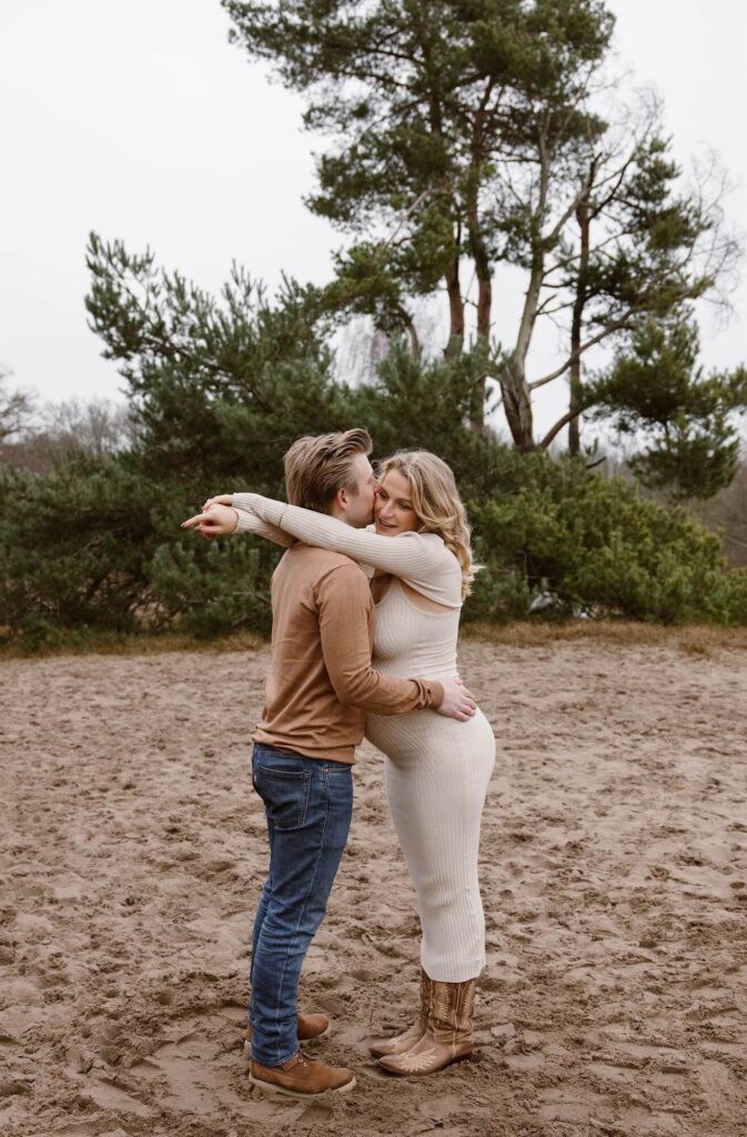 Zwangere vrouw in beige jurk en cowboylaarzen knuffelt man in bruine t-shirt en jeansbroek tijdens zwangerschapsshoot op heide