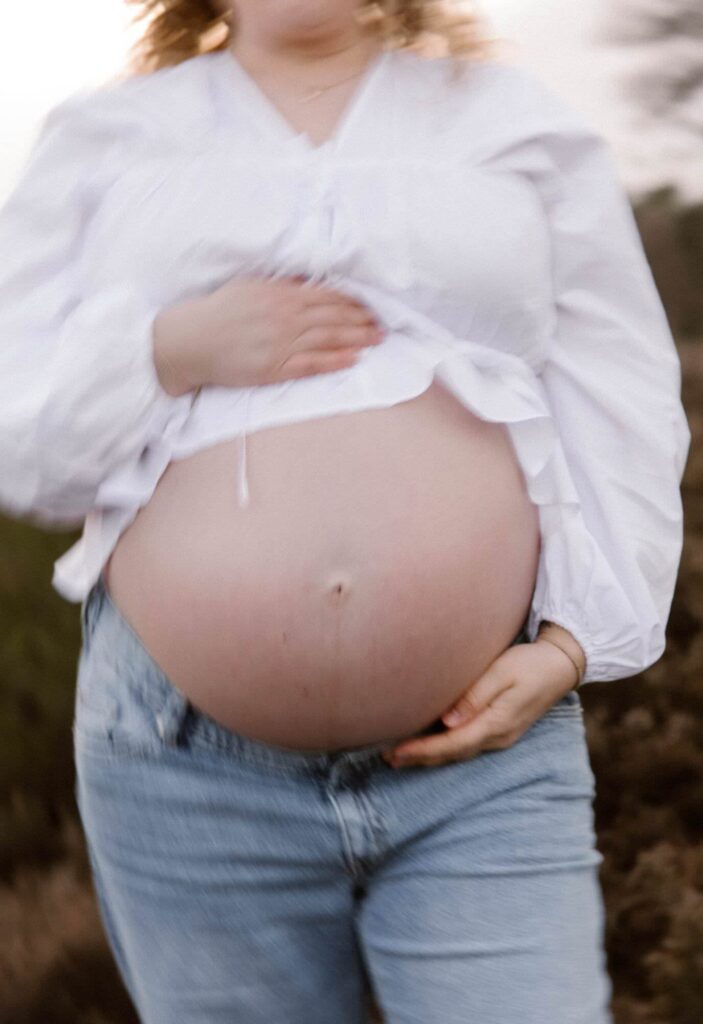 Zwangere vrouw poseert met handen op haar buik en draagt witte blouse met blauwe jeansbroek