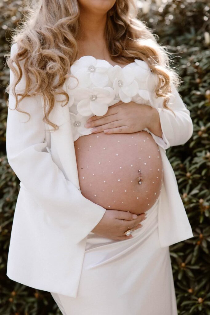 Zwangere vrouw in witte top met bloemen, witte blazer en witte rok houdt haar handen op haar buik met glitters tijdens zwangerschapsshoot