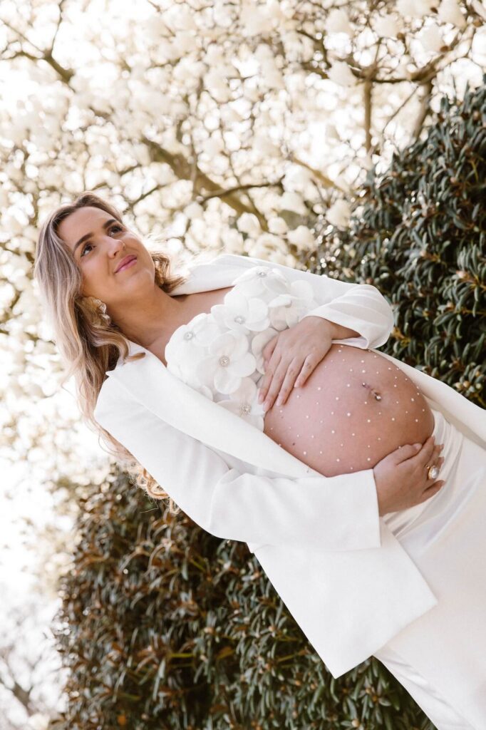 Zwangere vrouw in witte top met bloemen, witte blazer en witte rok houdt haar handen op haar buik met glitters tijdens zwangerschapsshoot