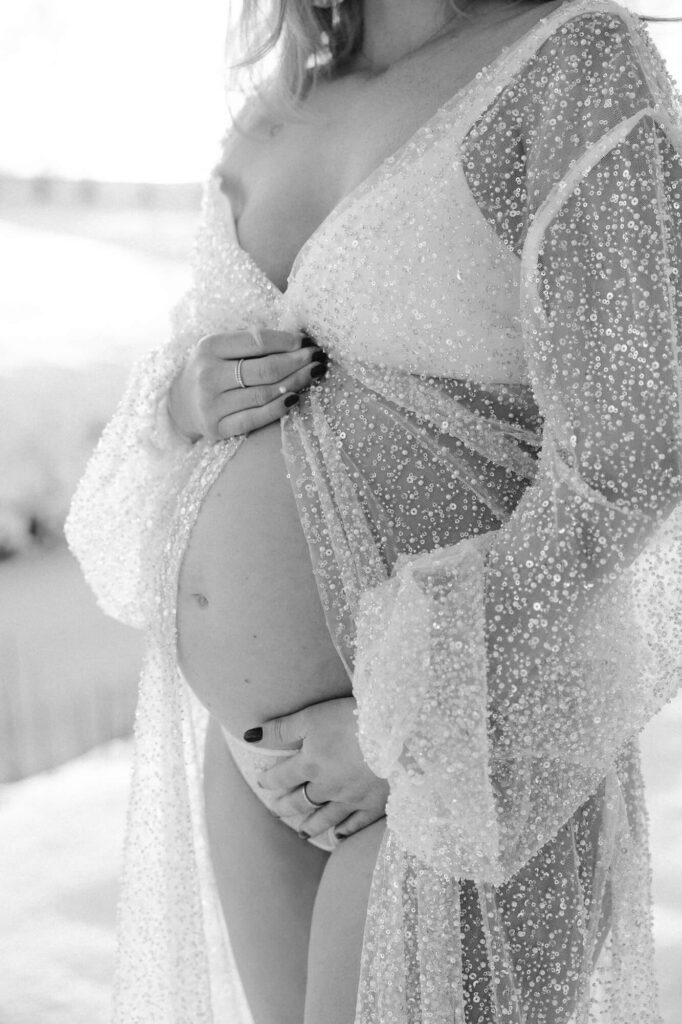 Zwart-wit close-up van vrouw in witte BH en doorzichtige kimono met glitters tijdens een zwangerschapsshoot in de sneeuw